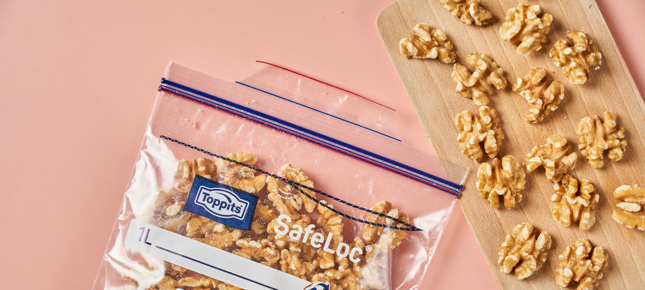Toppits SafeLoc Beutel gefüllt mit Walnusskernen liegt neben Holzbrett auf rosa Fläche.