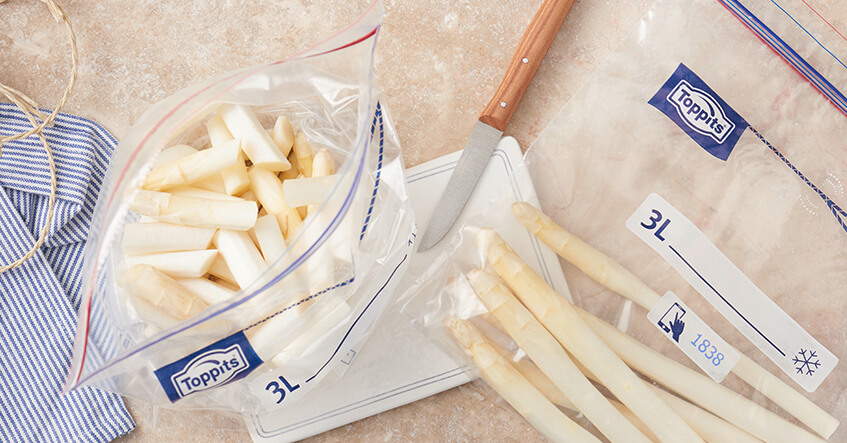 Weiße Spargelstücke in Toppits Gefrierbeutel auf Brett mit Messer und weiteren Beuteln.