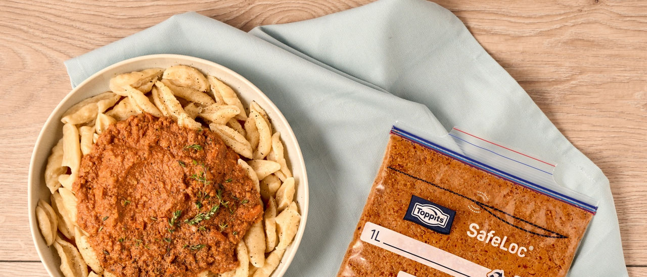 Teller mit Nudeln und Hackfleischsoße steht auf einem hellen Holztisch, daneben ein verschlossener Toppits SafeLoc Gefrierbeutel mit eingefrorenem Hackfleisch