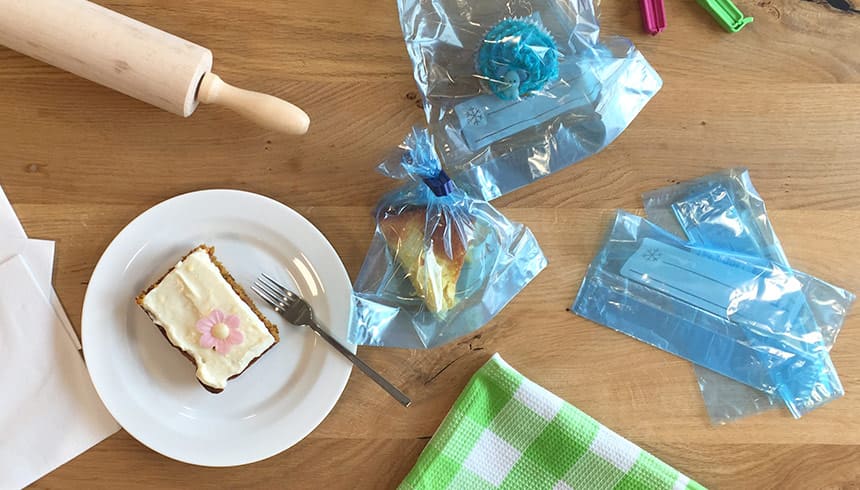 Gebäck auf einem Teller mit Gabel, daneben verpackte Muffins in Toppits Gefierbeutel , ein Nudelholz und ein grünes Geschirrtuch auf einem Holztisch 