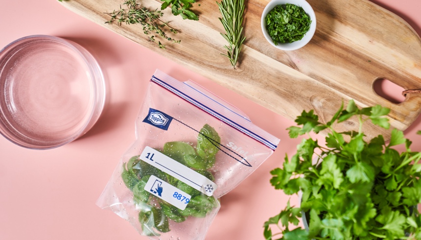Toppits Gefrierbeutel mit frischen Kräutern liegt auf einem rosa Untergrund neben einem Holzbrett mit weiteren Kräutern, einer Schale und frischen Kräutern im Vordergrund 